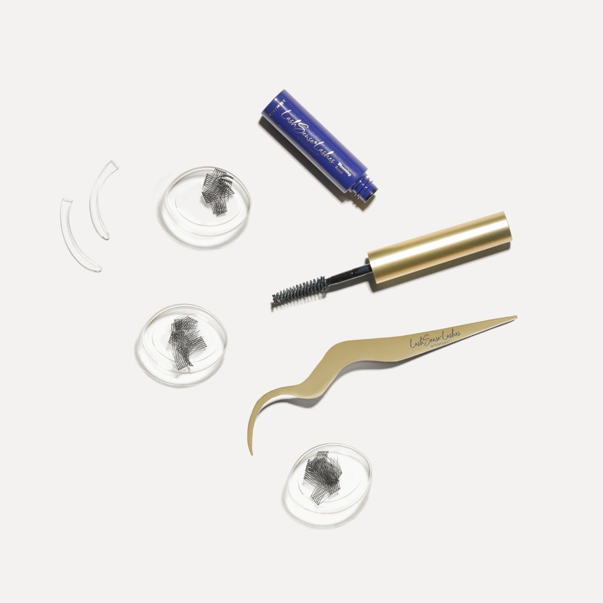 LashSense® Lashes Bloom Box by SeneGence: Black lash clusters in three clear round trays beside a blue adhesive tube, a gold-capped spoolie brush, two clear curved applicator guides and curved gold tweezers.