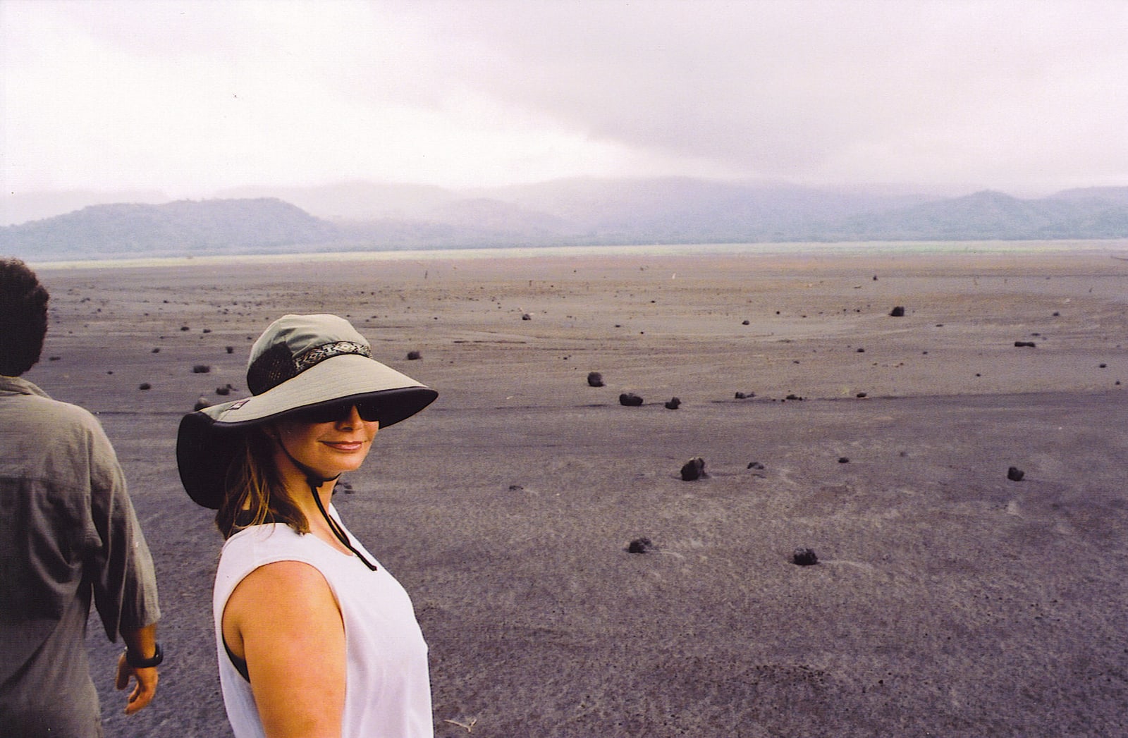 Joni Rogers-Kante in a broad-brimmed hat and sunglasses on a wide plain of dark volcanic ash in Vanuatu, misty hills behind her.